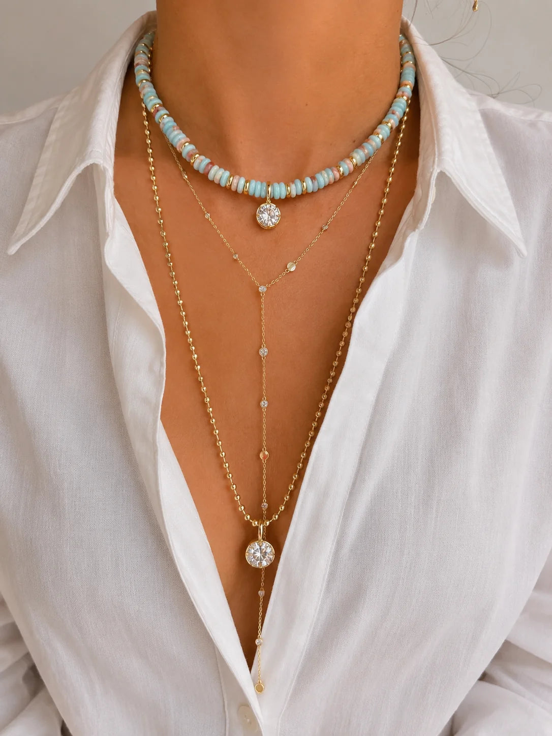 A close-up of a person wearing layered necklaces over a white collared shirt. The jewelry includes a choker with light blue and white beads, a gold Y-necklace with clear crystal-like round charms, and a longer gold chain.