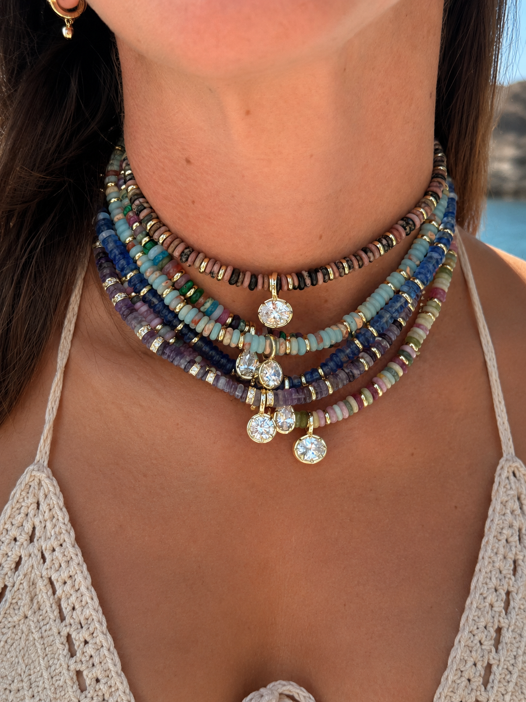A close-up of a woman wearing a multi-strand beaded choker necklace with gold-toned metal and clear rhinestone-like charms, photographed outdoors near water.